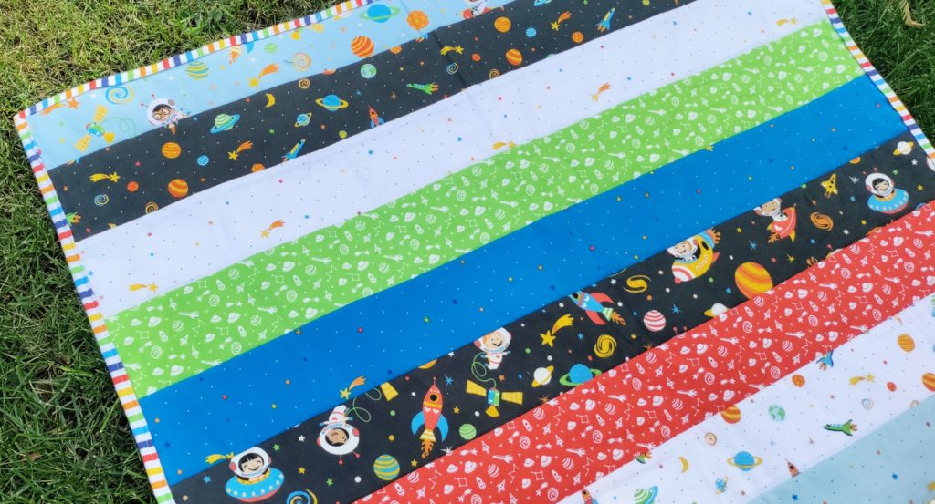 A quilt laying on the grass made with wide strips of the Blast Off fabric collection
