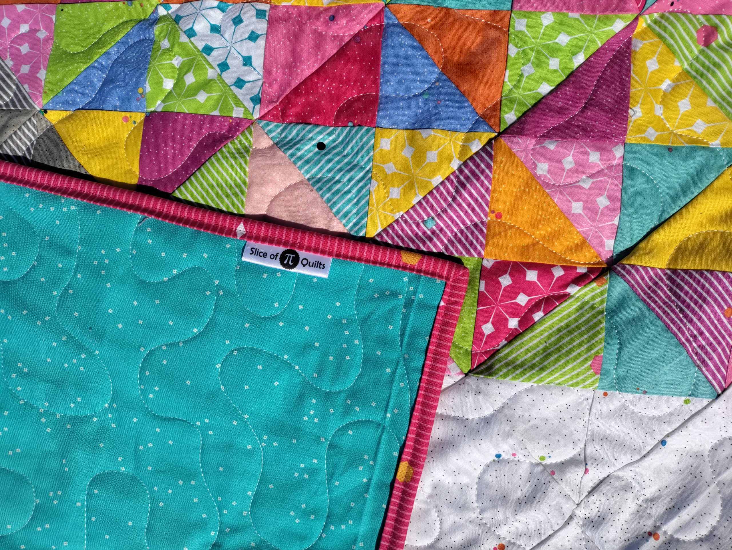 A satin quilt label sewn into the binding of the Flutterfly butterfly quilt. Aqua colored fabric is on the back of the quilt, and pink striped fabric is on the binding.