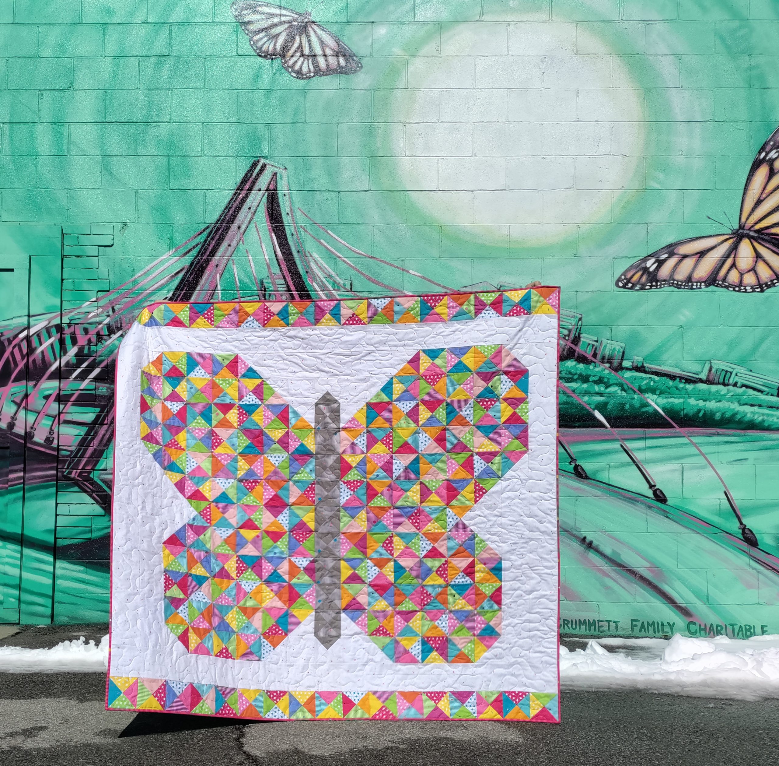 The Flutterfly butterfly quilt made with brightly colored fabric triangles of the Colour Wall collection by Riley Blake Designs in front of a green mural with a sun and Monarch butterflies.