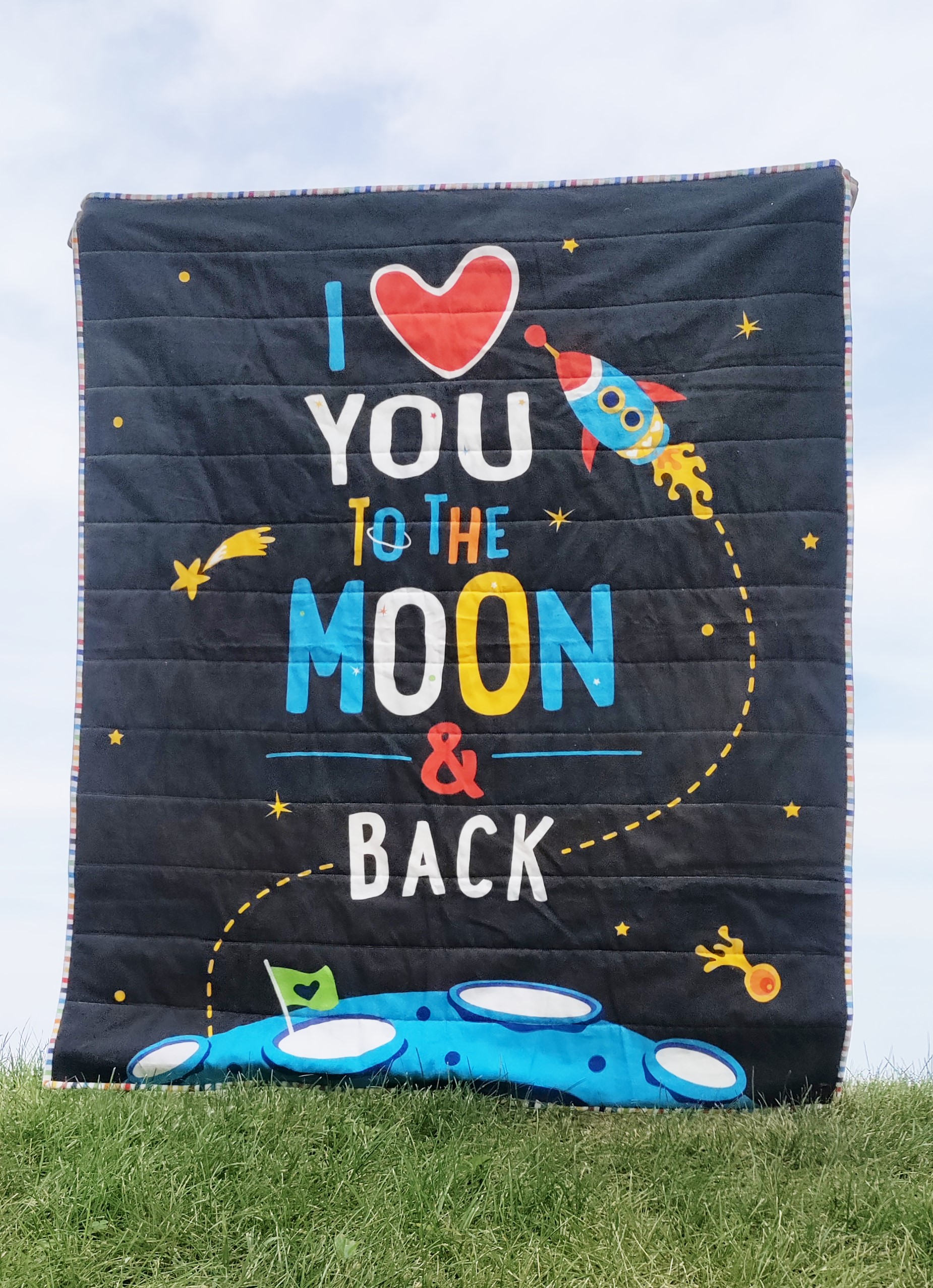 A quilt with the words "I love you to the moon and back" being held up by a young boy against a light colored sky