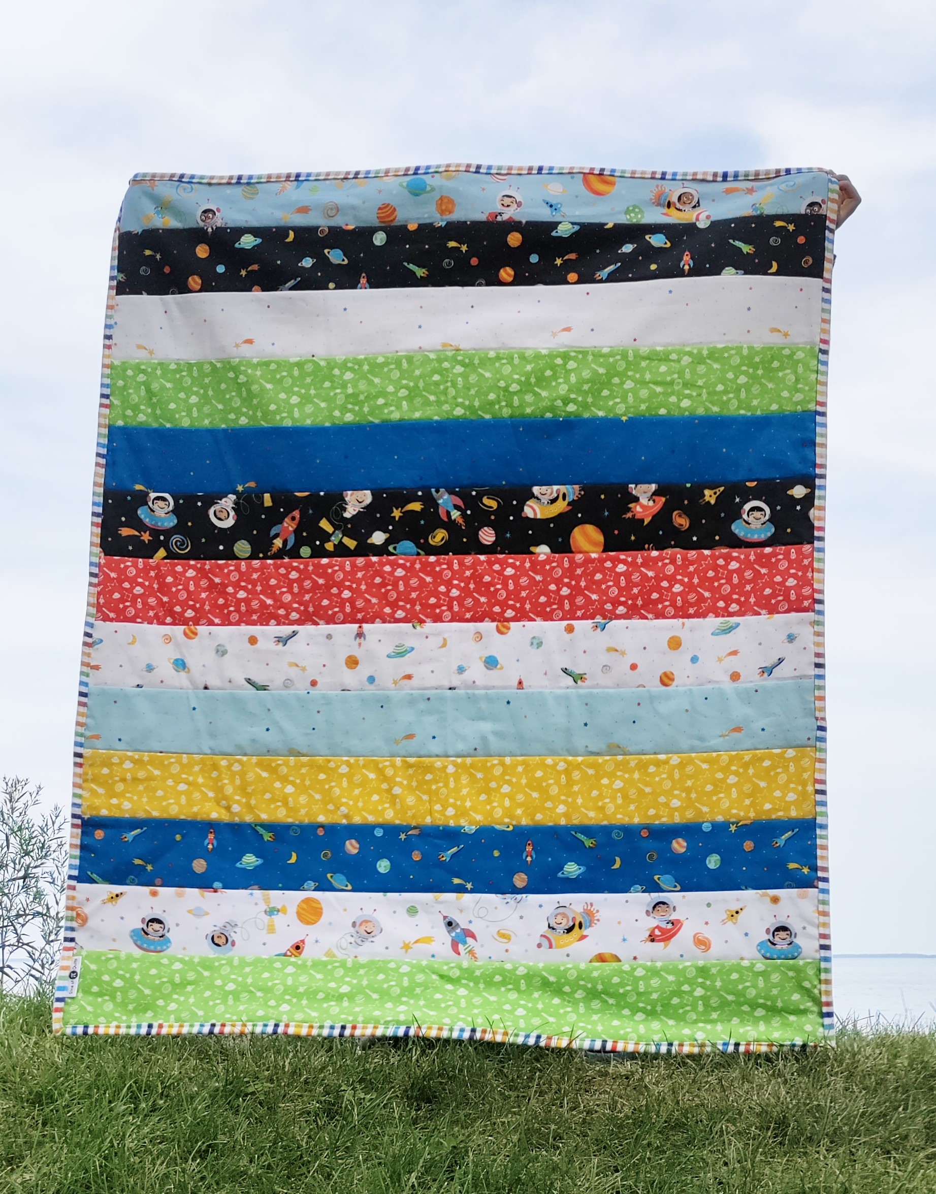 A quilt made from wide fabric strips being held up by a young boy against a light colored sky
